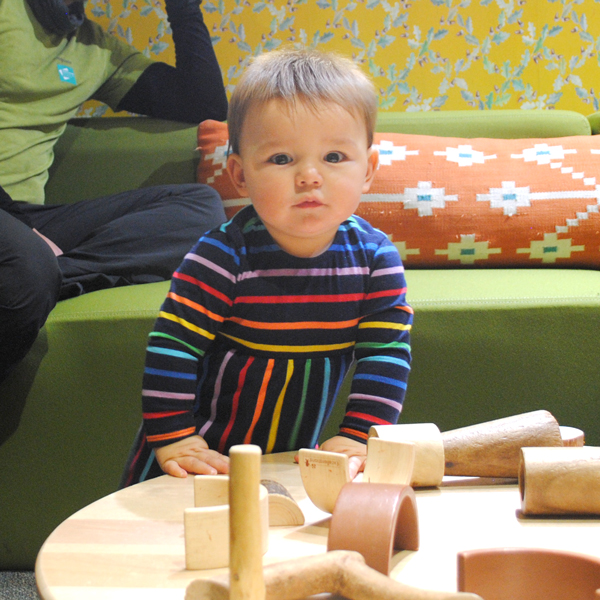 A baby pushes up on a wooden table covered in wooden blocks and manipulatives, while an adult reclines in the background on a couch