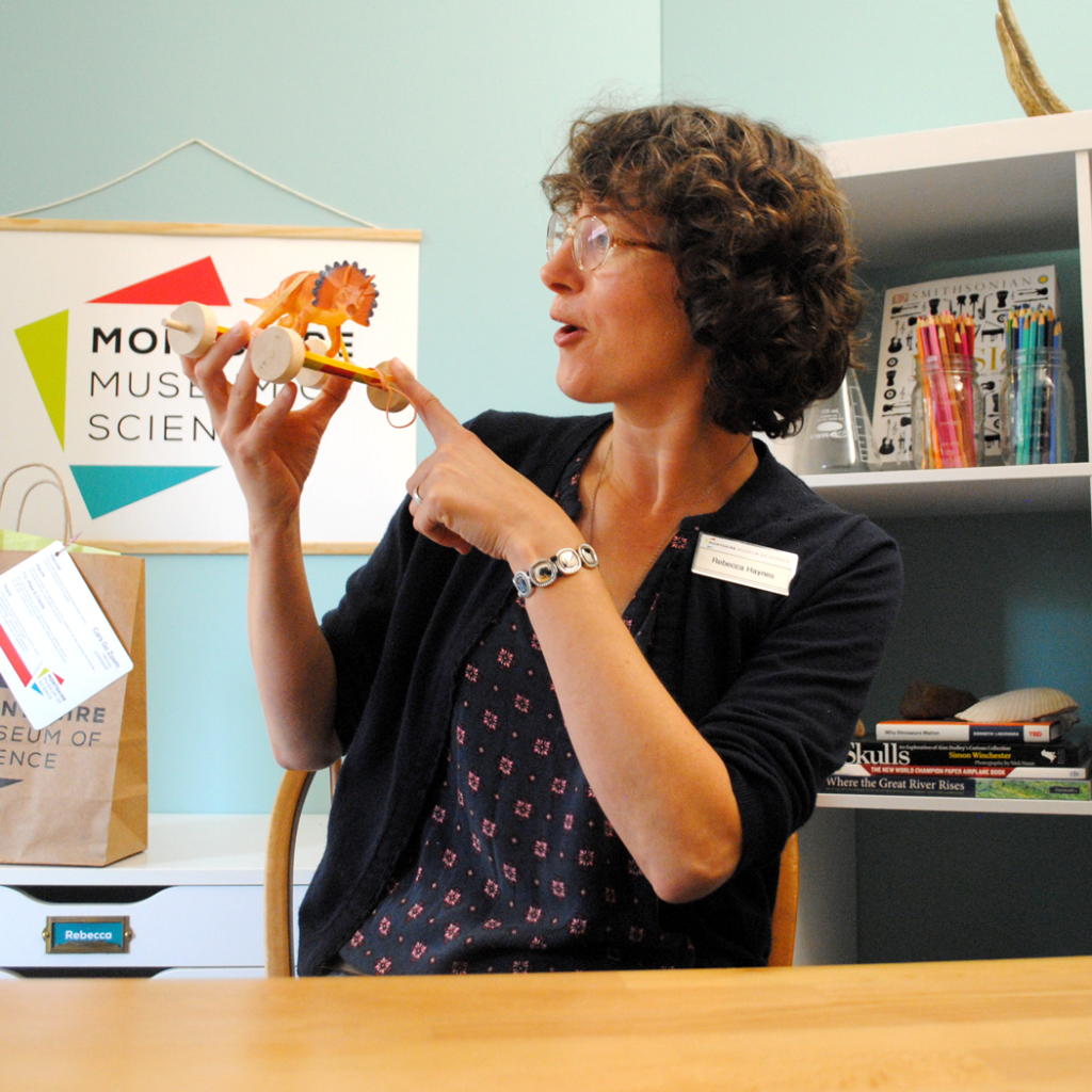 Montshire educator pointing at a specimen while seated at a desk with a Montshire logo behind.