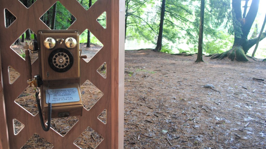 An old fashioned wooden phone with handset is set against wooden latticework, behind which you can see a woodland forest floor and the bases of tree trunks.