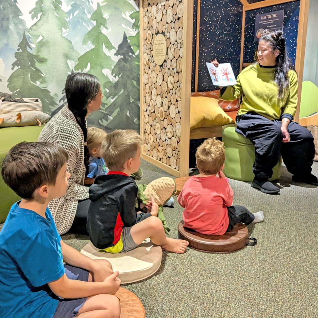 A Montshire educator reads a story to young visitors and caregivers seated in WonderWoods
