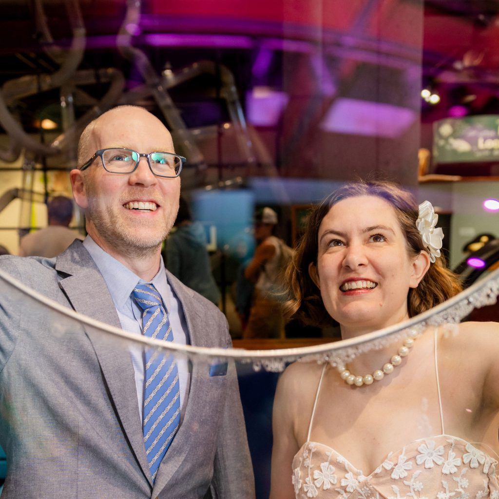 A smiling couple in wedding clothes lift a giant bubble frame at the Montshire Museum of Science